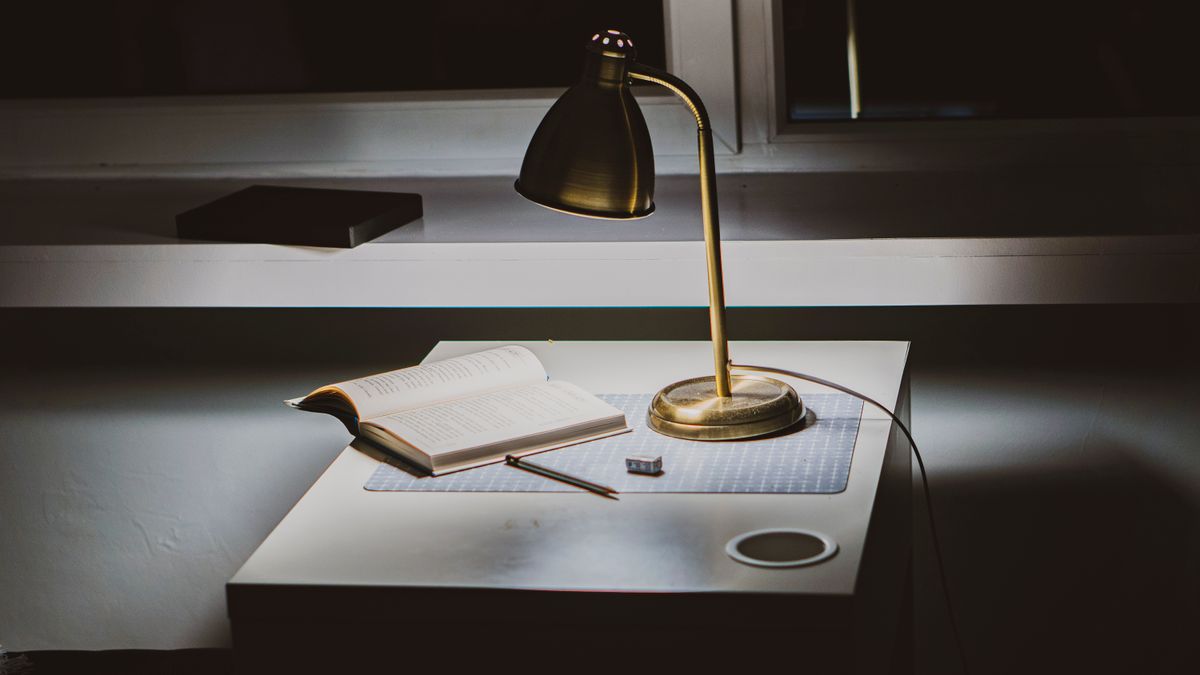 Brass desk lamp lighting an open book and pencil on a desk in the evening