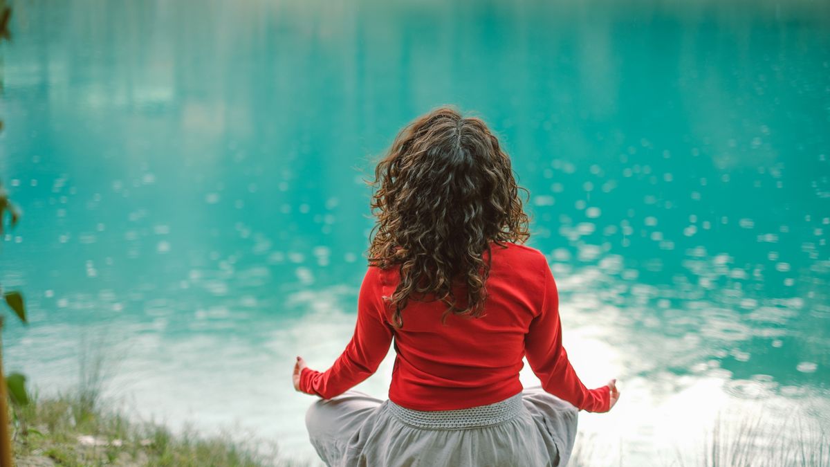 Woman in a red top meditating cross-legged beside a calm turquoise lake