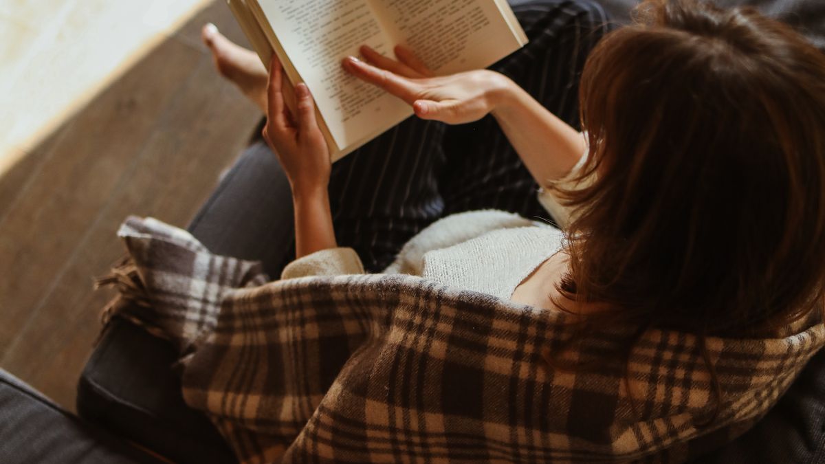 Woman wrapped in a plaid blanket reading a book alone on a sofa