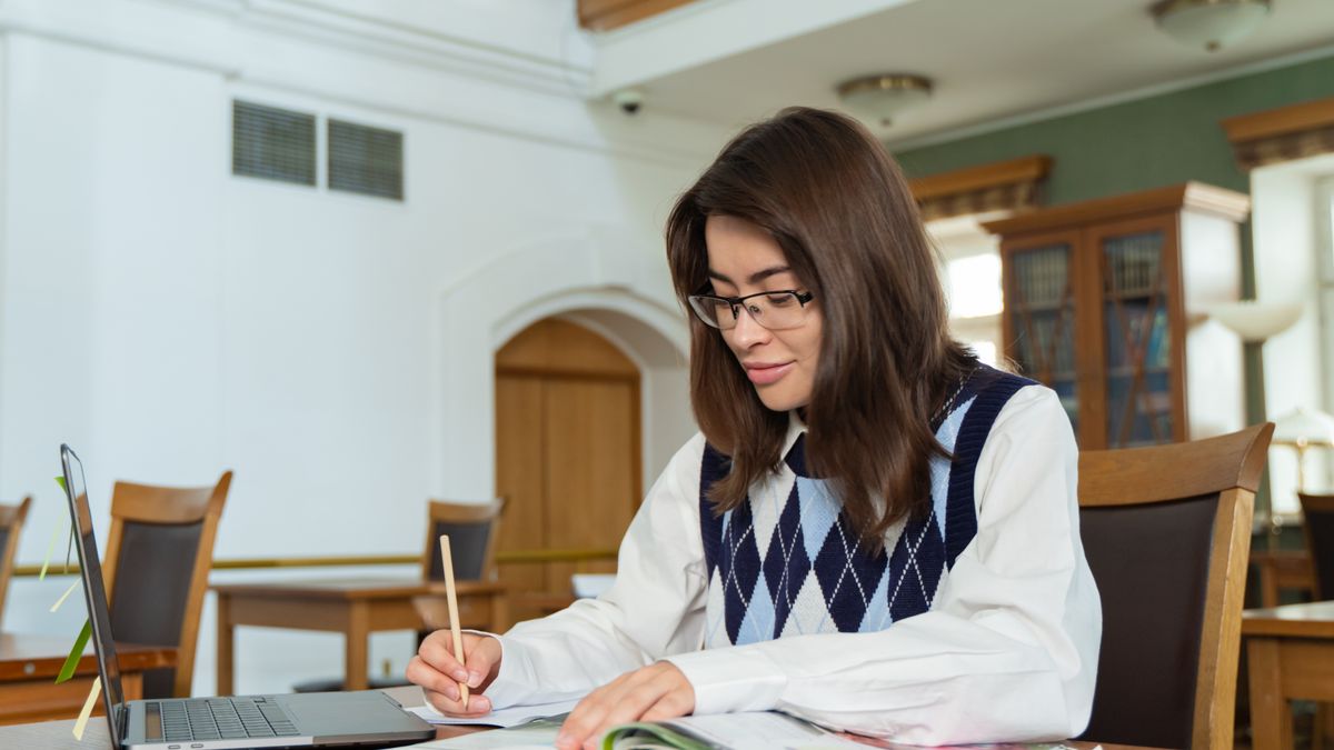 Student in glasses taking notes from an open book at a library table