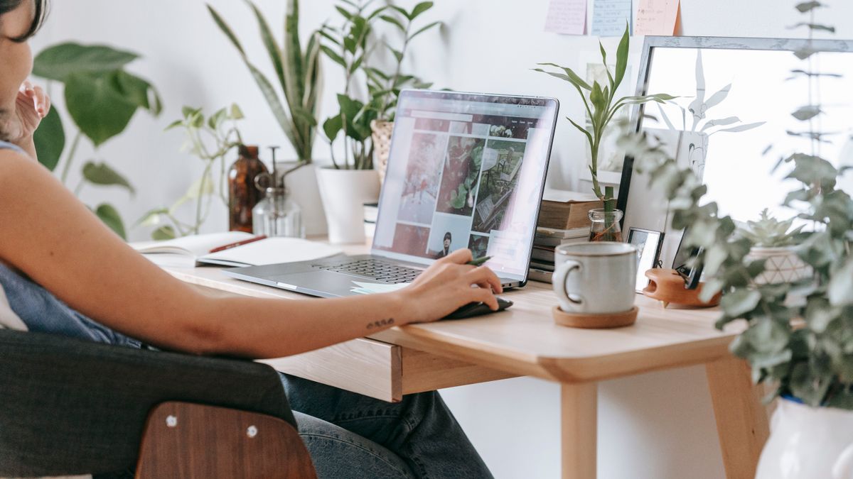 Woman working on a laptop at a wooden home desk surrounded by plants